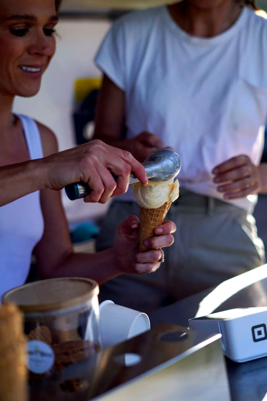 Hands hand-scooping a generous scoop of pale Jersey vanilla into a waffle cone